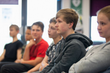 Teenage boy by sitting in a row of teenagers looking towards the front of the room