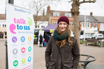 Young lady wearing a hat and coat standing next to a Healthwatch banner