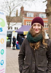 Young lady wearing a hat and coat standing next to a Healthwatch banner