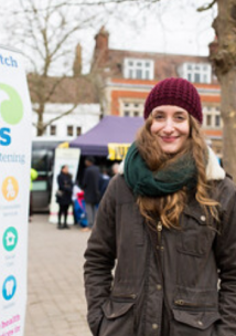 Young lady wearing a hat and coat standing next to a Healthwatch banner